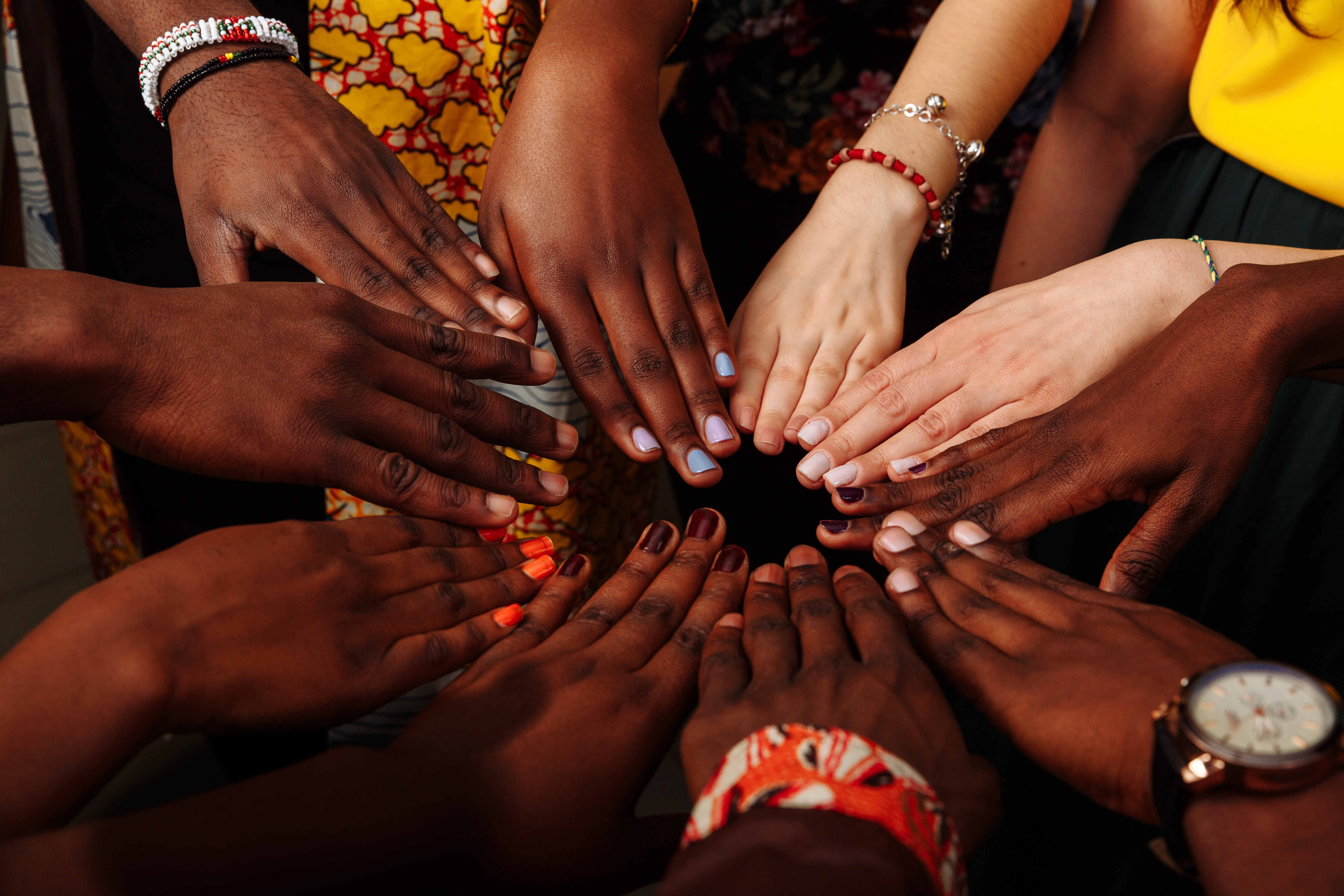 Hands of women with varied skin tones placed together in a circle, bracelets and a wristwatch visible.