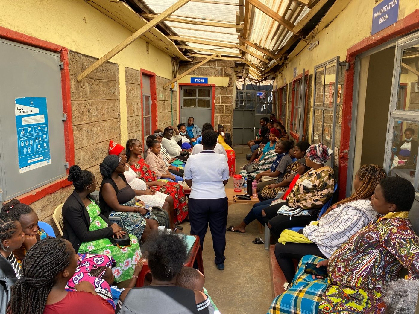 A midwife in uniform speaks to a group of mothers seated in a community clinic waiting area.