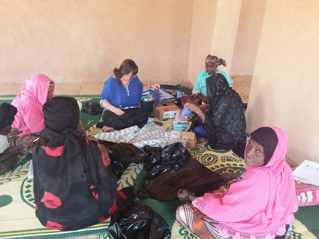 A midwife in a patterned headscarf works alongside a colleague in scrubs at a hospital bedside, with a young girl seated nearby.