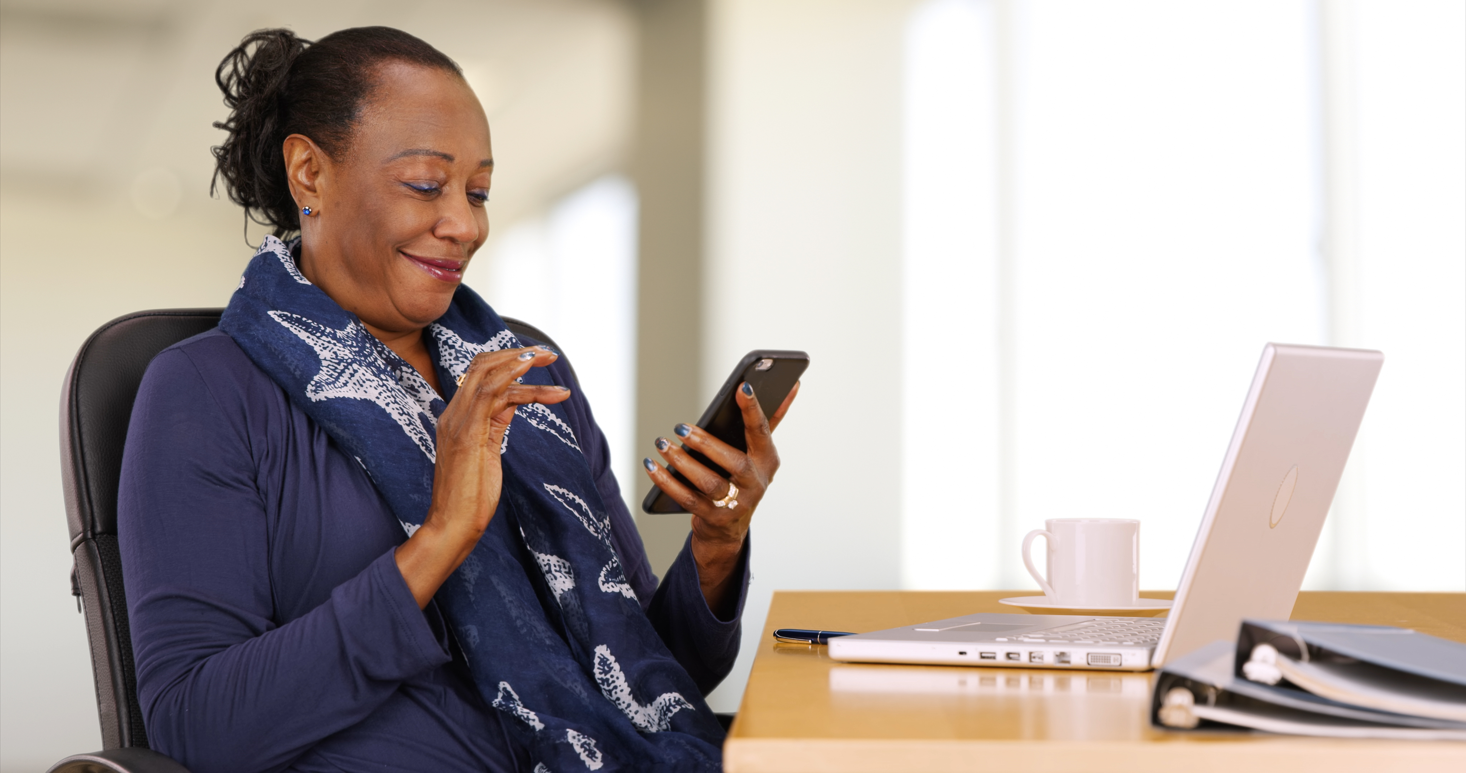 A midwife smiles as she taps on her smartphone at a desk, with a laptop and notebook beside her.