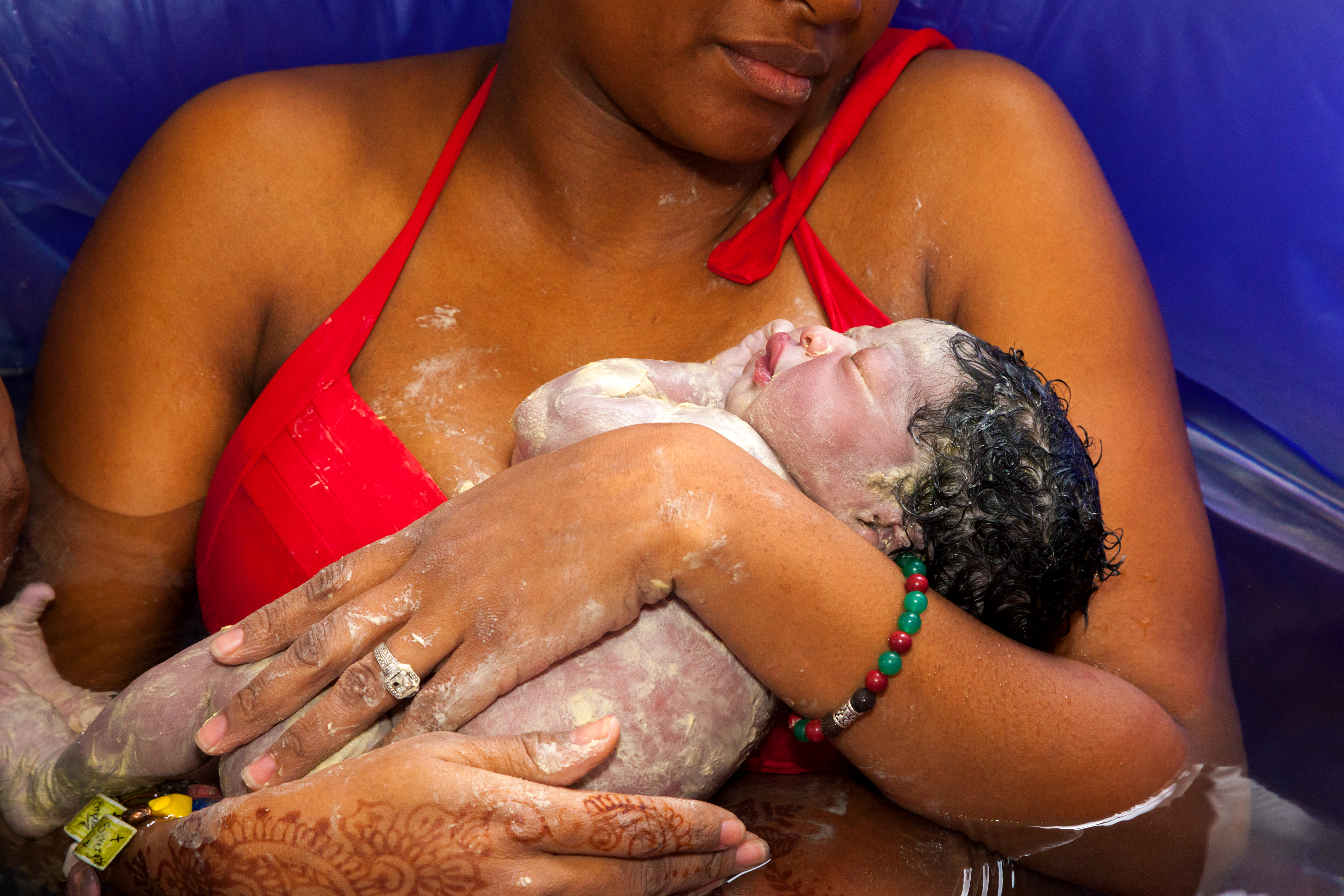 A mother in a red top cradles her newborn moments after a water birth, henna decorating her hand.
