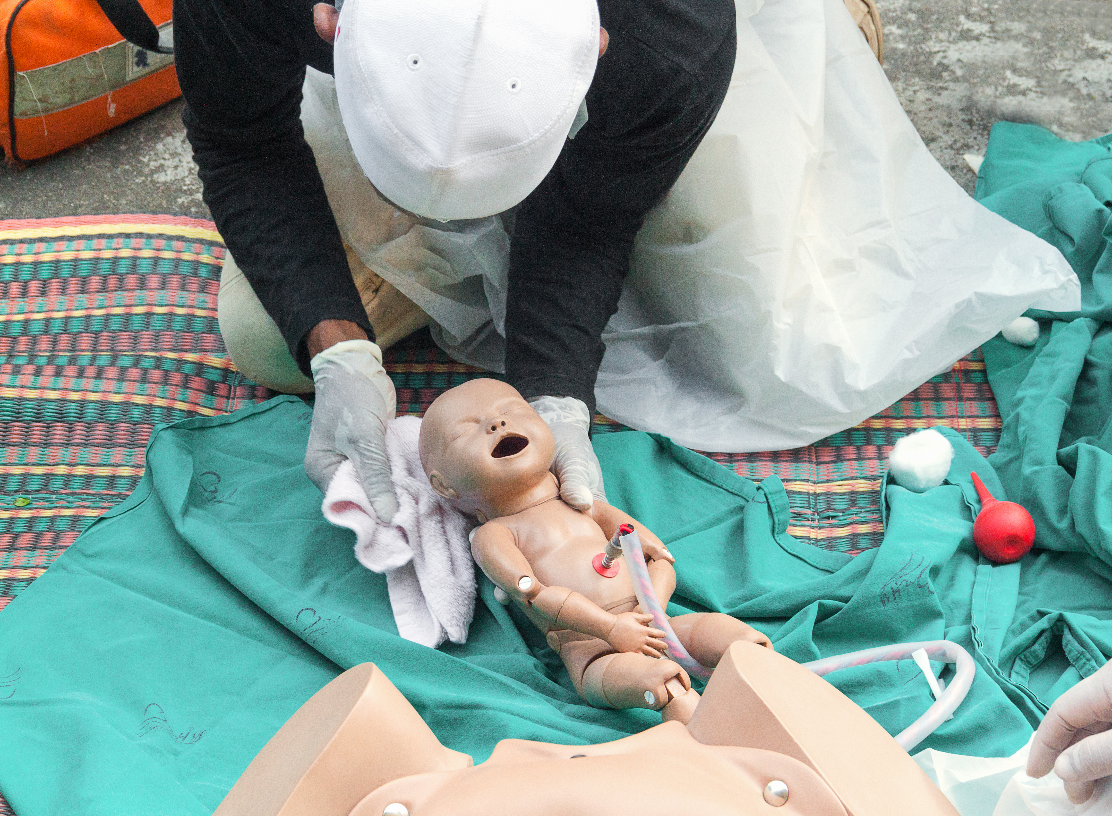 A trainee in gloves practices newborn resuscitation on an infant manikin, with bulb syringe, cord clamp, and emergency kit laid out alongside.
