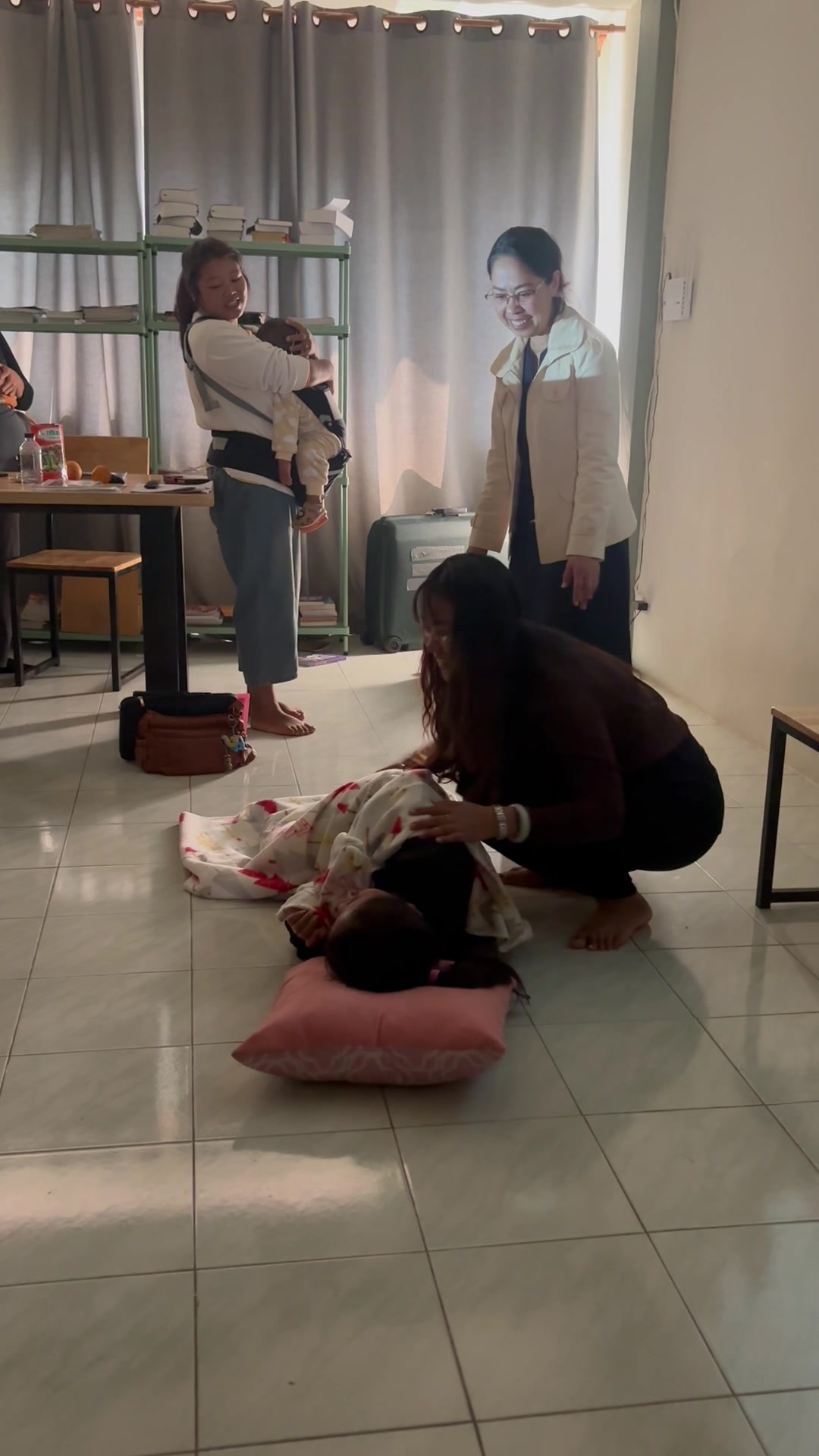A trainer kneels beside a participant lying on a pillow on the floor, demonstrating a skill with cloth, while a colleague in a white jacket smiles as she observes.